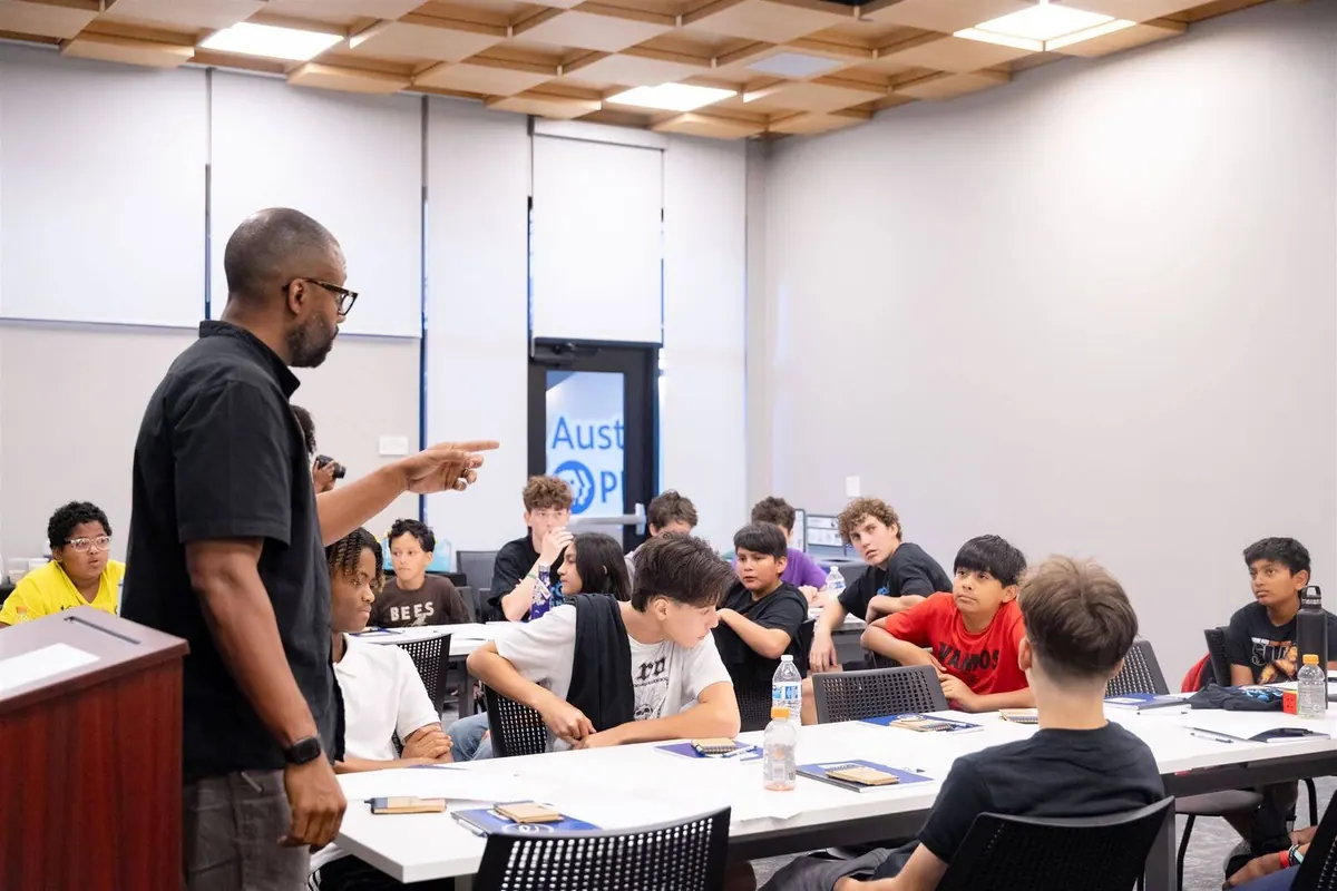 Bavu Blakes speaks to a group of young learners seated around tables in a classroom.